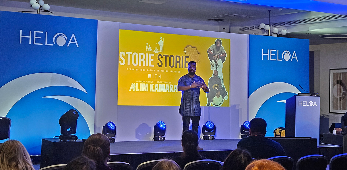 Alim Kamara stands on stage with an animated expression, the screen behind is yellow with the Storie Storie logo