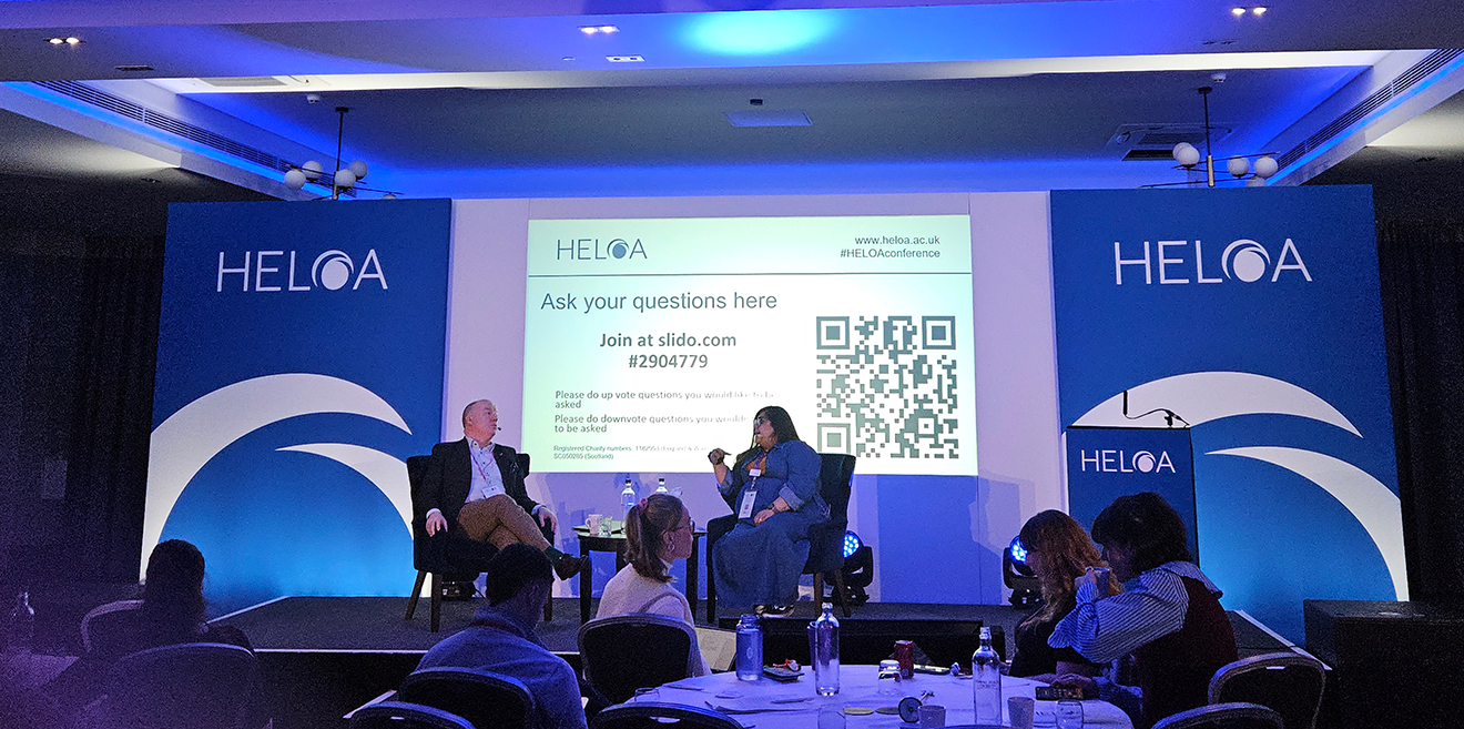 Reena Littlehales interviewing Professor Ken Sloane on a blue HELOA branded stage